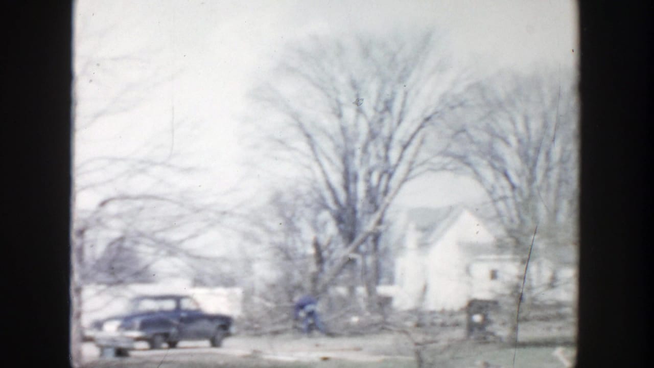 1956 Grand Rapids residential winter view with a parked blue sedan, snow on the ground, bare trees, and houses beyond dark window framing.