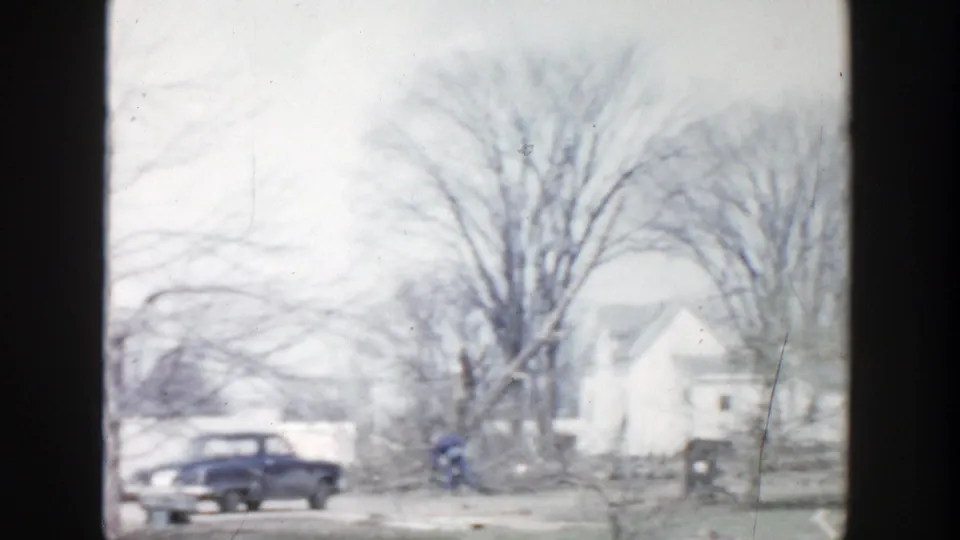 1956 Grand Rapids residential winter view with a parked blue sedan, snow on the ground, bare trees, and houses beyond dark window framing.