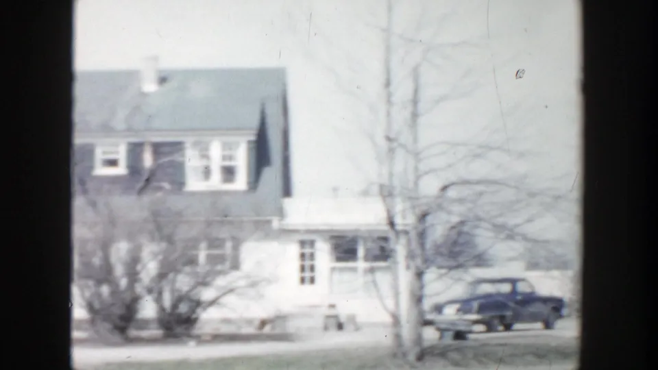 Residential Grand Rapids winter scene with a white house, larger gray-roofed home, bare trees, and a dark mid-century sedan near the street.