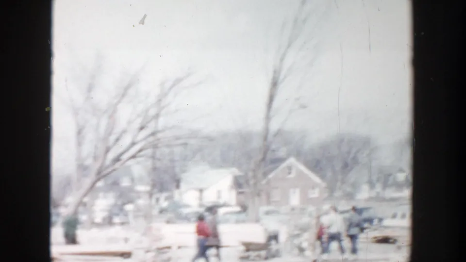 1956 Grand Rapids neighborhood view showing bundled pedestrians near benches or low street fixtures, with parked cars and houses in snow.
