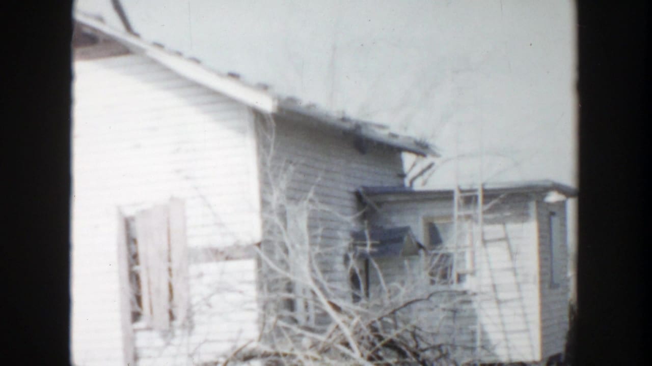 White house exterior with broken windows, roof damage, bare branches, and dark film borders.