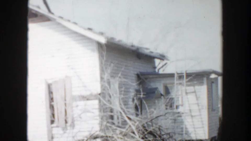 1956 Grand Rapids 8mm frame showing a white clapboard house with damaged roof edges, broken windows, bare branches, and a side structure.