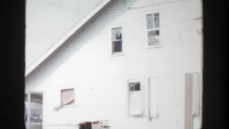 1956 Grand Rapids 8mm frame with an angled view of the white house side, showing sash windows, a lower opening, roofline, and overexposed siding.