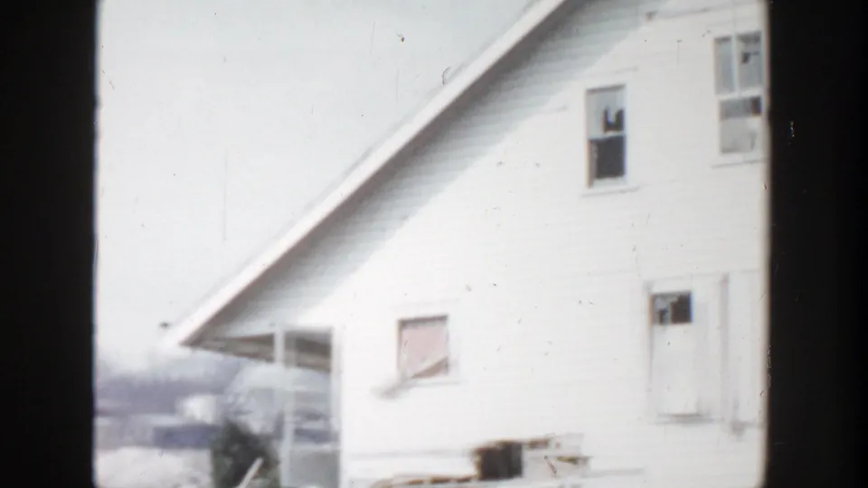 1956 Grand Rapids home-movie frame showing a pale clapboard house exterior with a boarded window and soft 8mm exposure.