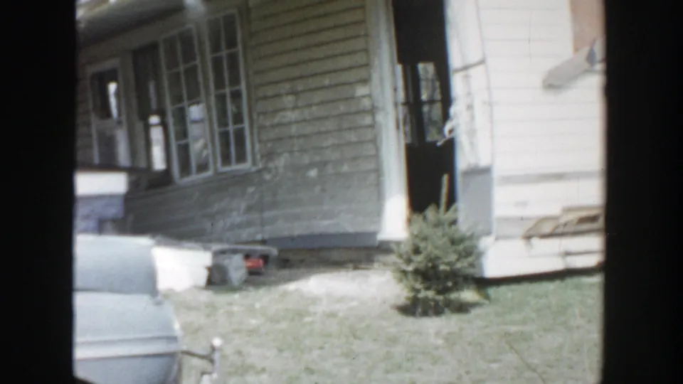 1956 Grand Rapids frame showing a weathered clapboard wall, porch windows, an open dark doorway, lawn clutter, and part of a car.