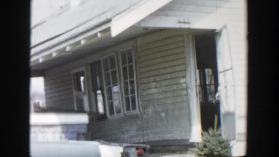 Grand Rapids 1956 home-movie frame with a close exterior view of porch windows, clapboard siding, open doorway, and roof eave.