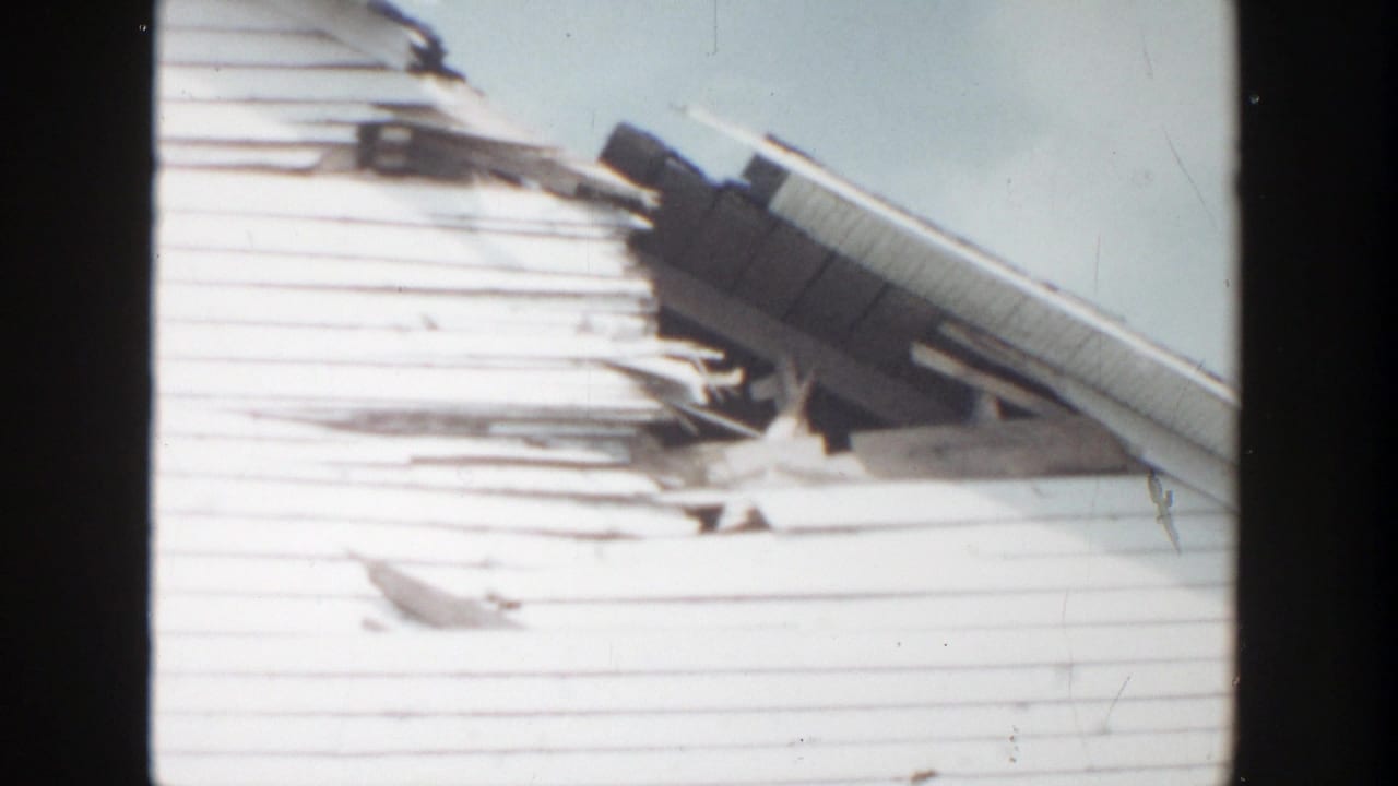 1956 Grand Rapids home-movie frame showing torn white siding, bent roof material, and exposed structure above a light brick wall.