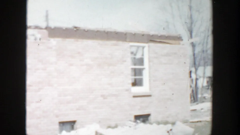 1956 Grand Rapids home exterior showing a pale brick wall, window, snow near the foundation, and damaged roof area at the top edge.