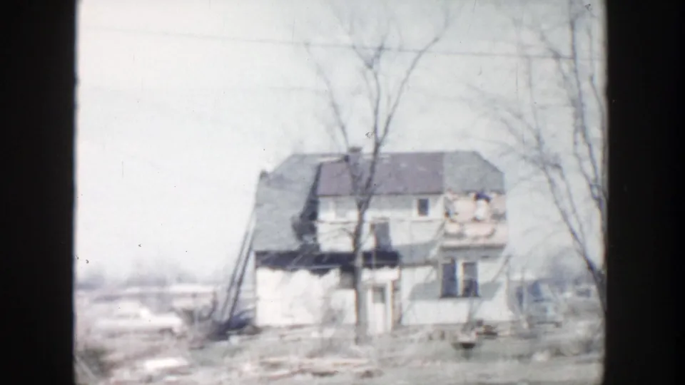 1956 Grand Rapids-area frame showing a damaged house, bare tree branches, scattered debris, and snow patches in a rural yard.