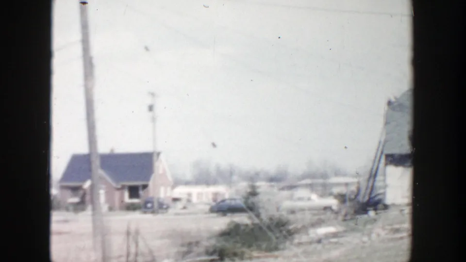 1956 Grand Rapids-area 8mm view across a winter roadside with modest houses, utility poles, parked cars, and damaged siding at frame edge.