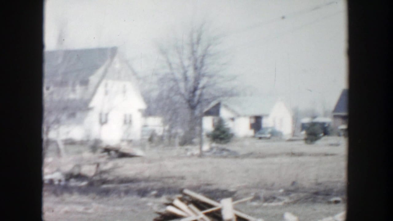 Damaged residential yard with a white house, bare tree, outbuildings, and scattered debris.