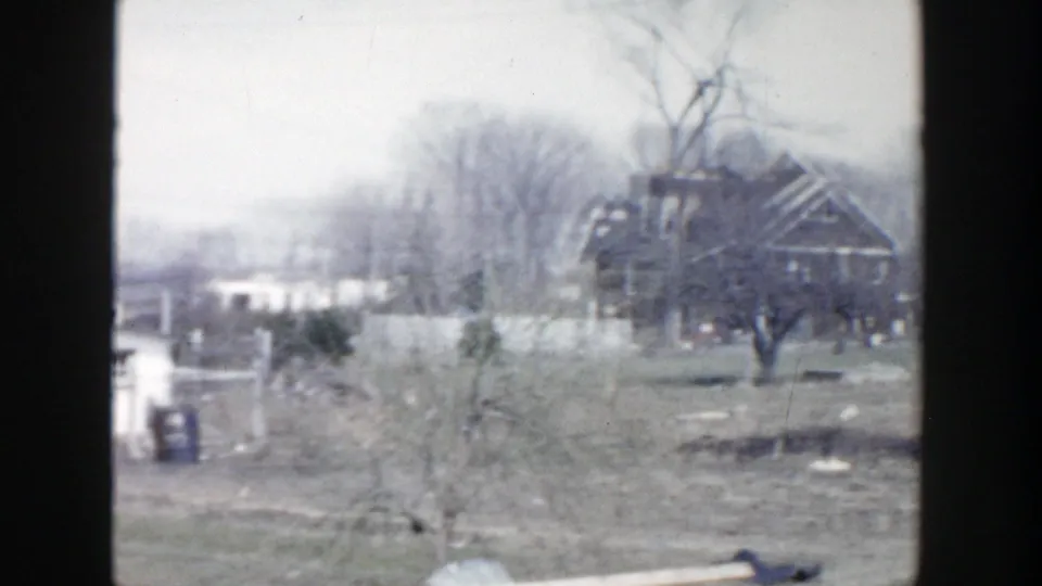 1956 Grand Rapids frame showing a damaged brick house, small foreground structure, bare trees, and scattered debris.