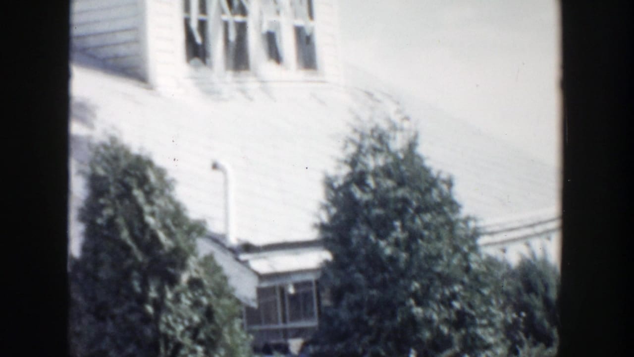 1956 Grand Rapids home-movie frame showing a white clapboard house exterior with windows and a large evergreen shrub in front.