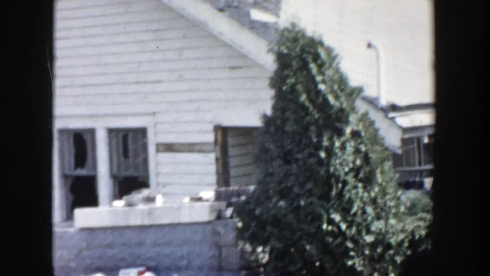 1956 Grand Rapids frame of a white house exterior with broken windows, a small side structure, and a prominent evergreen shrub.