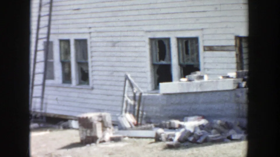 1956 Grand Rapids residential exterior showing broken windows, debris piles, a ladder, and steps beside a white clapboard house.