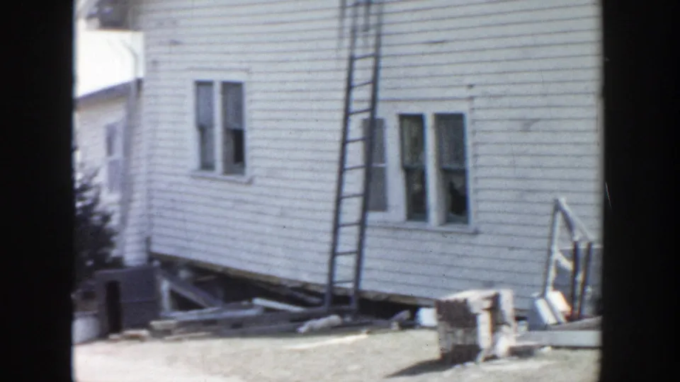 1956 Grand Rapids home-movie frame showing the side of a white clapboard house with a ladder, broken window, and scattered debris.