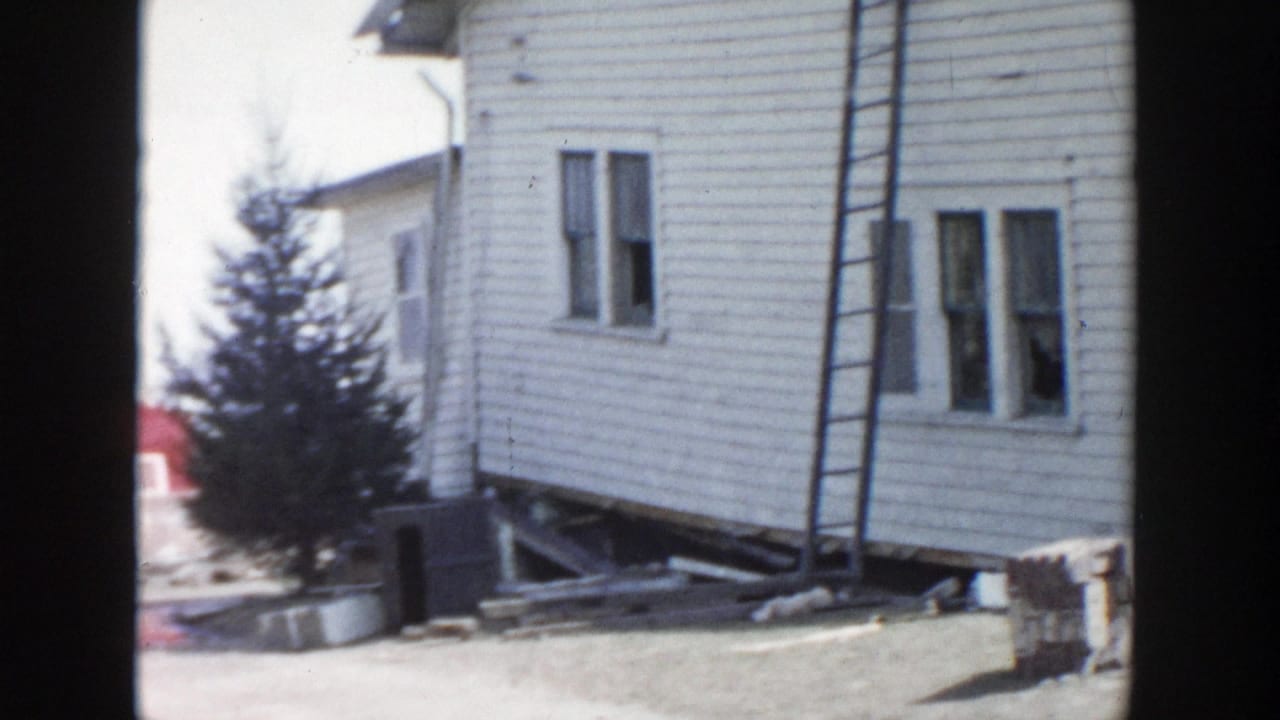 1956 Grand Rapids home-movie frame showing a white clapboard house elevated on supports with a ladder and yard debris nearby.