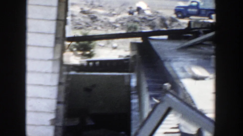 1956 Grand Rapids frame looking through dark foreground edges toward the raised house area, with a blue truck visible beyond the yard.