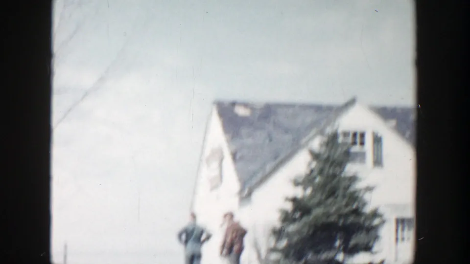 Two men stand outside a white house in Grand Rapids, Michigan, seen through soft 8mm home-movie framing.