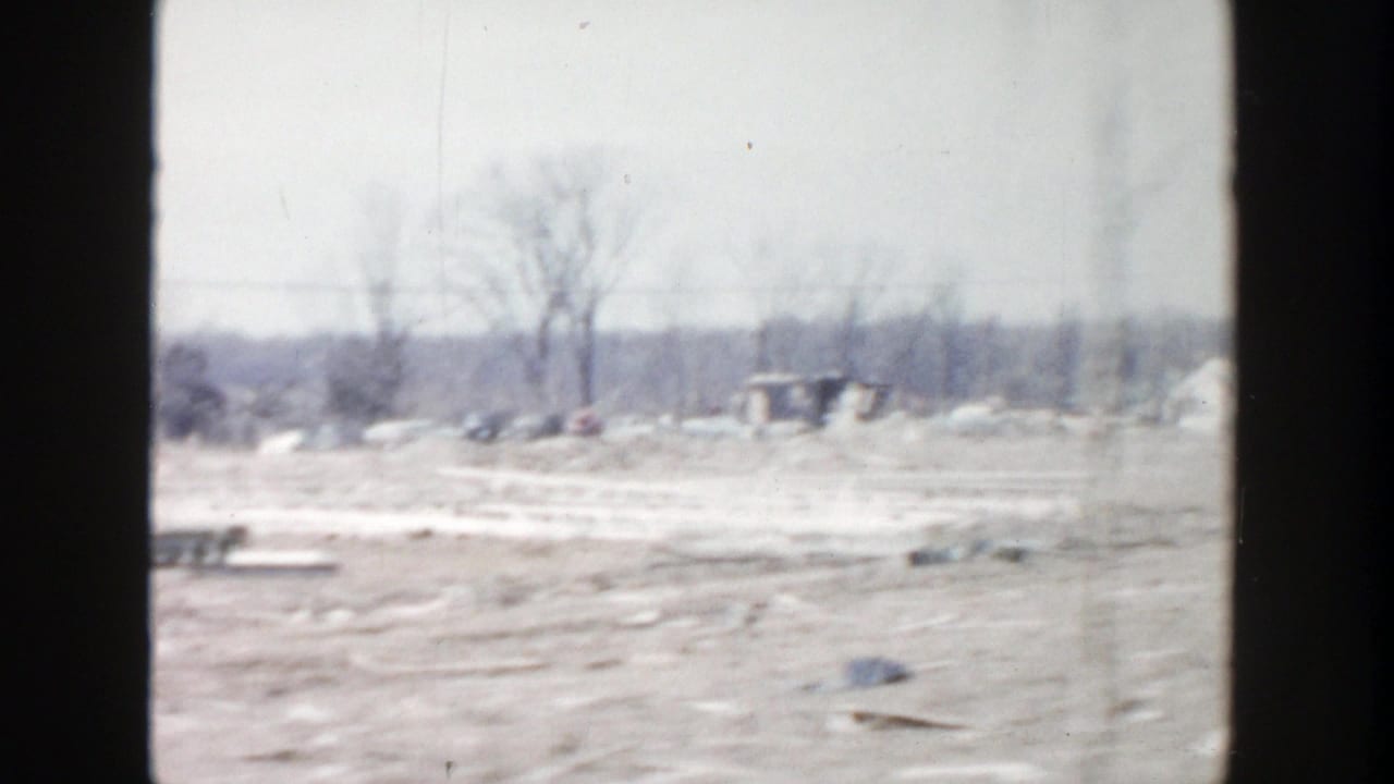 Muddy open ground with debris, bare trees, and distant low buildings under a pale sky.