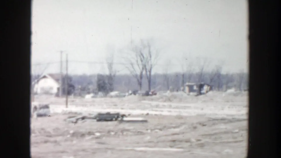 1956 Grand Rapids view of a damaged open area with utility poles, a small white house, bare trees, and scattered wreckage.