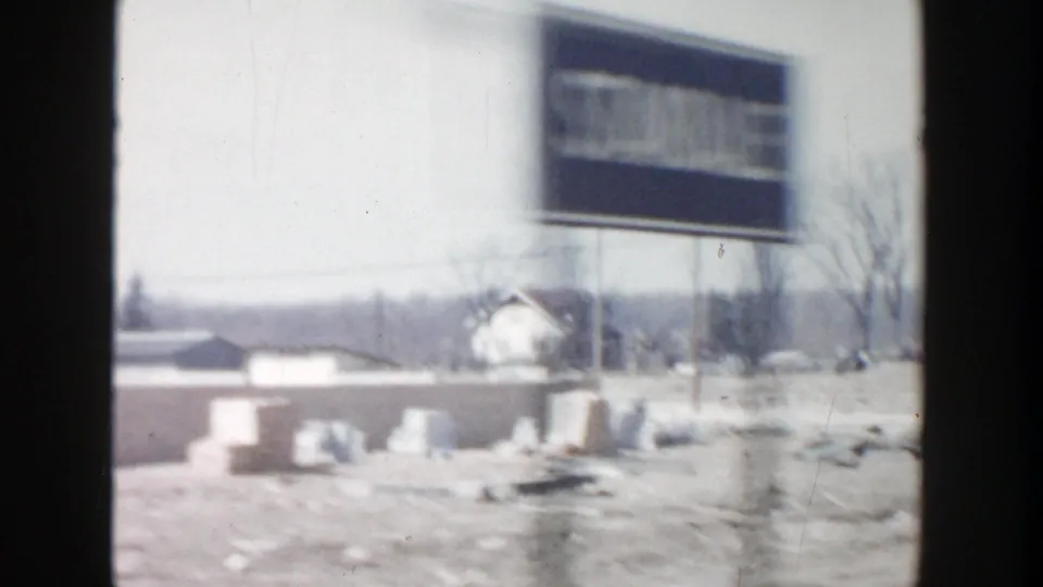 Drive-by 1956 Grand Rapids frame with a blurred roadside sign, low structures, scattered debris, and a pale car near the horizon.