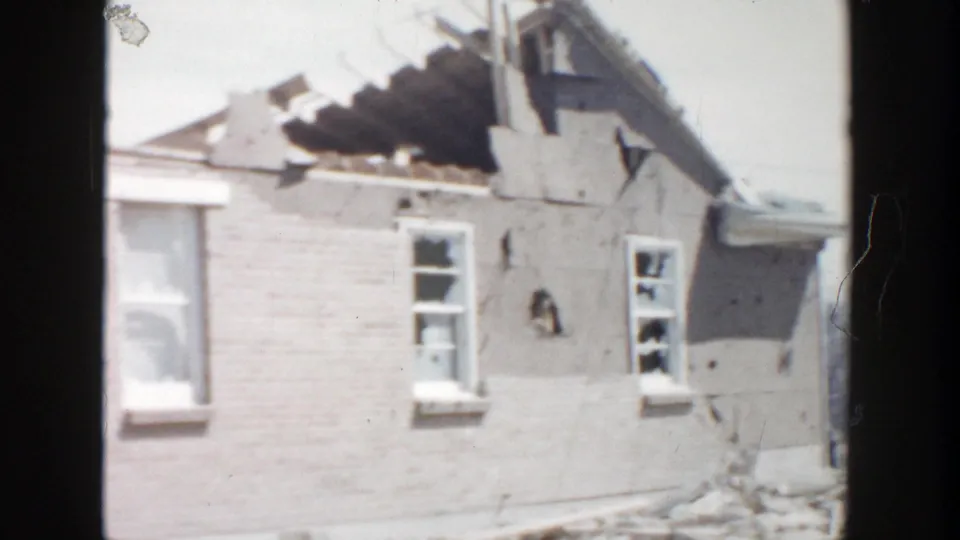 1956 Grand Rapids frame showing a damaged brick house with shattered windows, exposed roof structure, and debris below.