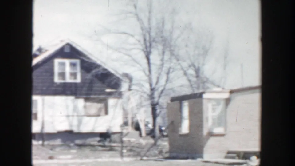 1956 Grand Rapids home-movie frame with neighboring houses, a bare tree, yard debris, and tiny distant people near the work area.