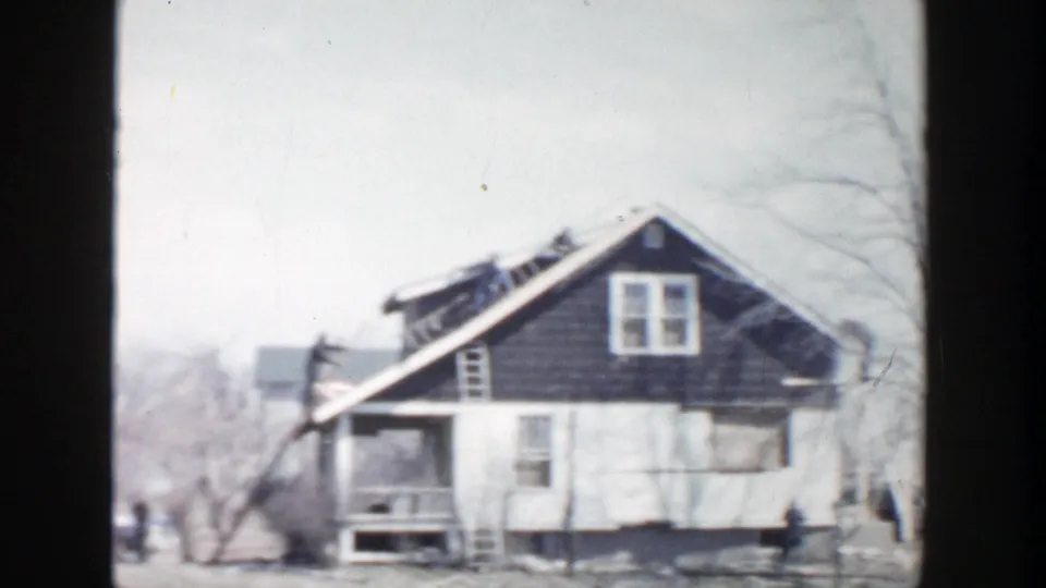1956 Grand Rapids frame showing a wood-sided house with roof damage, porch area, ladder, bare trees, and distant figures.