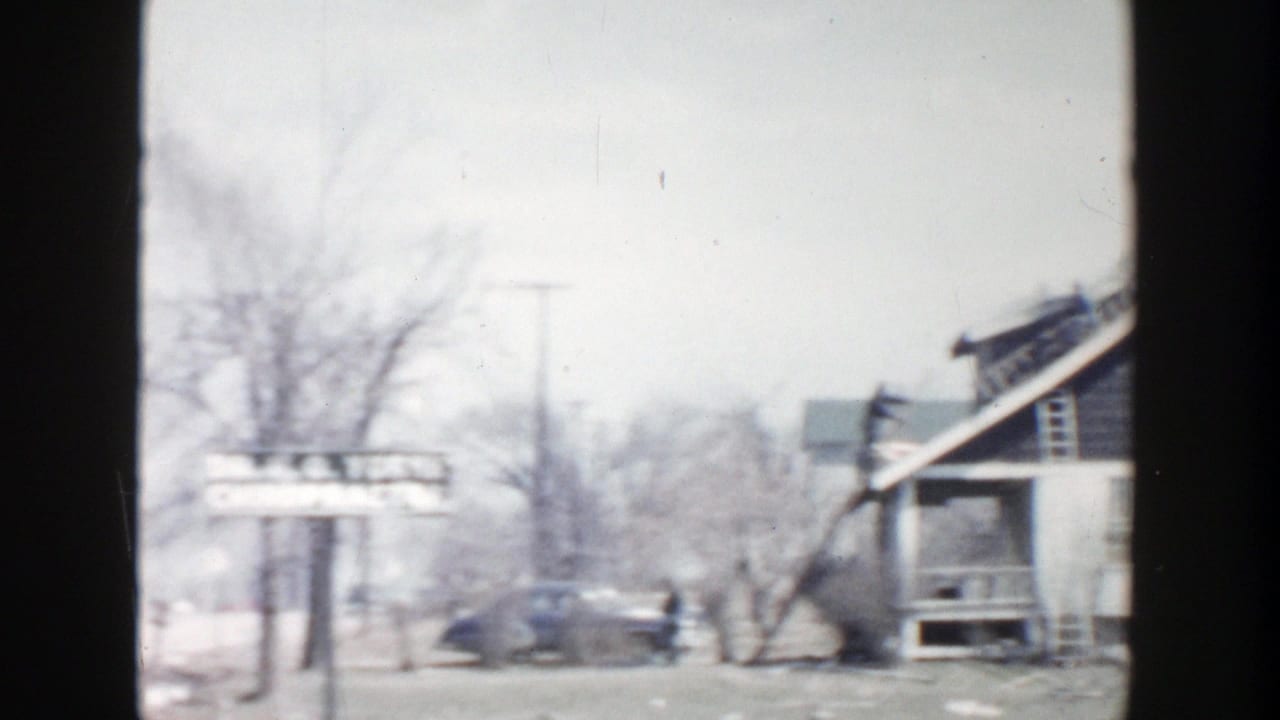 1956 Grand Rapids street view with older cars, bare trees, storefront facades, and a sign along the sidewalk.