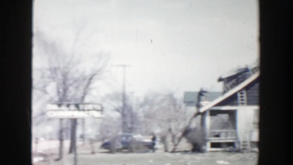 1956 Grand Rapids street view with older cars, bare trees, storefront facades, and a sign along the sidewalk.