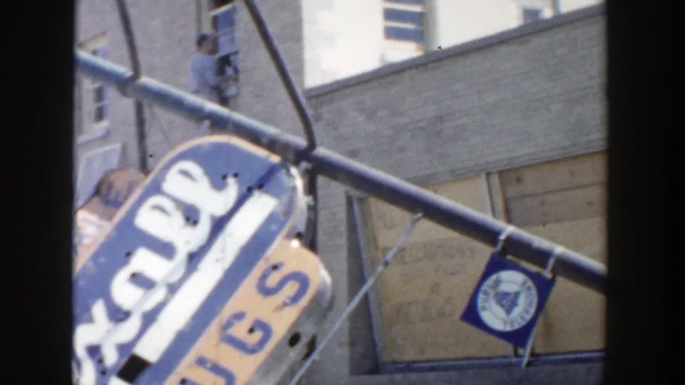 Damaged blue and orange Rexall Drugs sign hanging askew outside a Grand Rapids storefront in 1956.