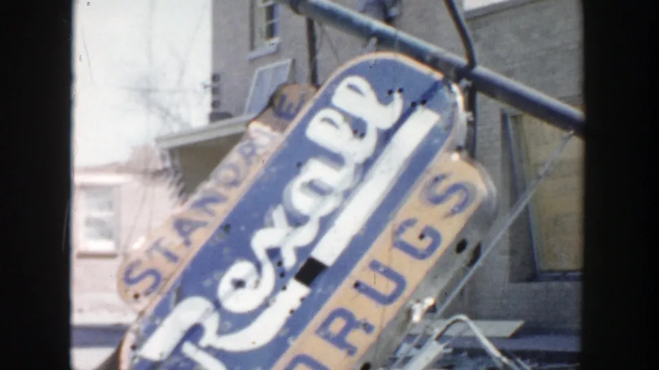 Close 1956 view of the damaged Rexall Drugs sign, with boarded storefront windows and nearby buildings behind it.