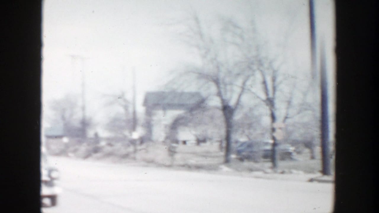 1956 8mm frame from Grand Rapids showing a snow-dusted roadside with bare trees, modest houses, and a forward-moving vehicle viewpoint.