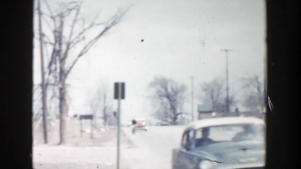 Grand Rapids winter road scene with a roadside sign, leafless trees, snow along the shoulder, and a mid-century car entering the frame.