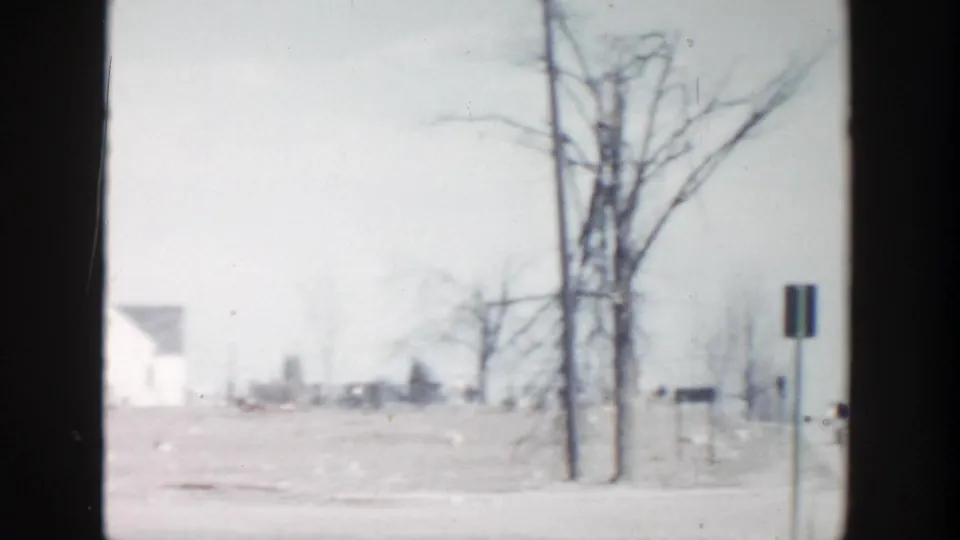 1956 home-movie frame showing a Grand Rapids roadside with snow, bare trees, a signpost, and distant modest houses.