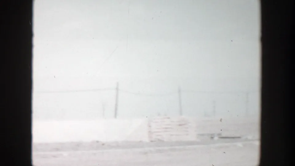 1956 Grand Rapids frame with a pale roadside structure, utility poles, and washed daylight in the background.