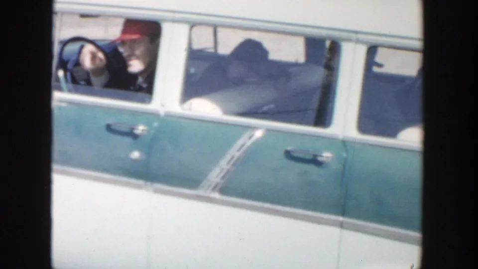 1956 Grand Rapids home-movie frame showing a blue and white station wagon with an adult driver in a red hat and another adult passenger visible.