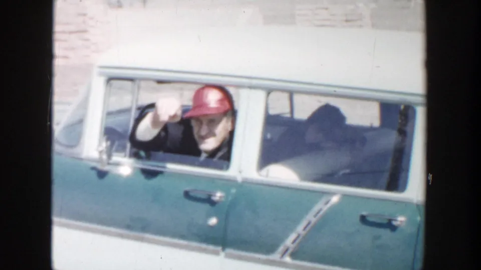 1956 Grand Rapids frame showing an adult man in a red hat and glasses waving from the driver window of a blue and white station wagon.