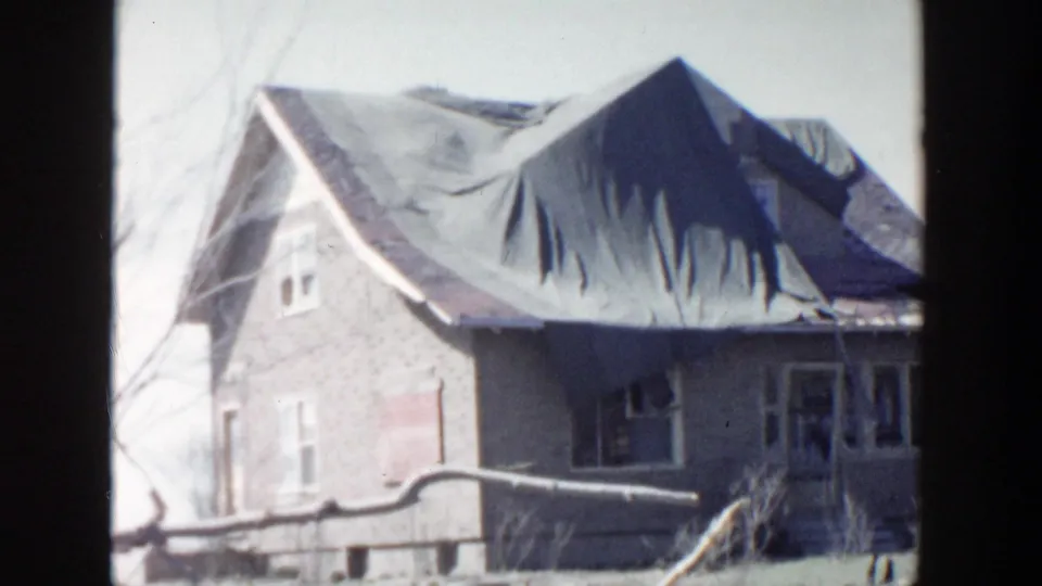 The damaged house exterior remains in view, with a large roof tarp, visible windows, and branches in the foreground.