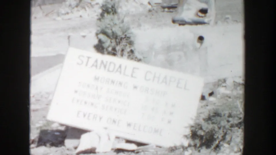 1956 Grand Rapids home-movie frame showing the Standale Chapel sign at an angle, with faint service text and rubble nearby.