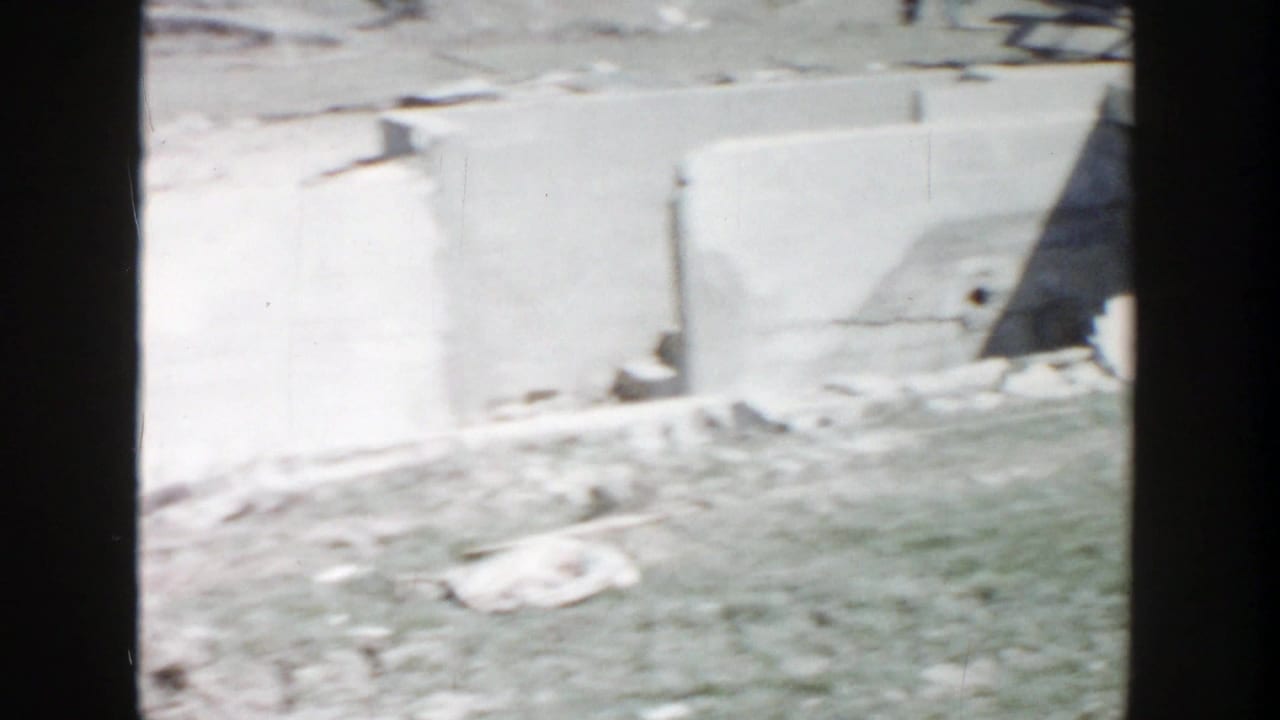 1956 Grand Rapids construction-site view with low concrete walls, loose materials, and open ground in soft 8mm focus.