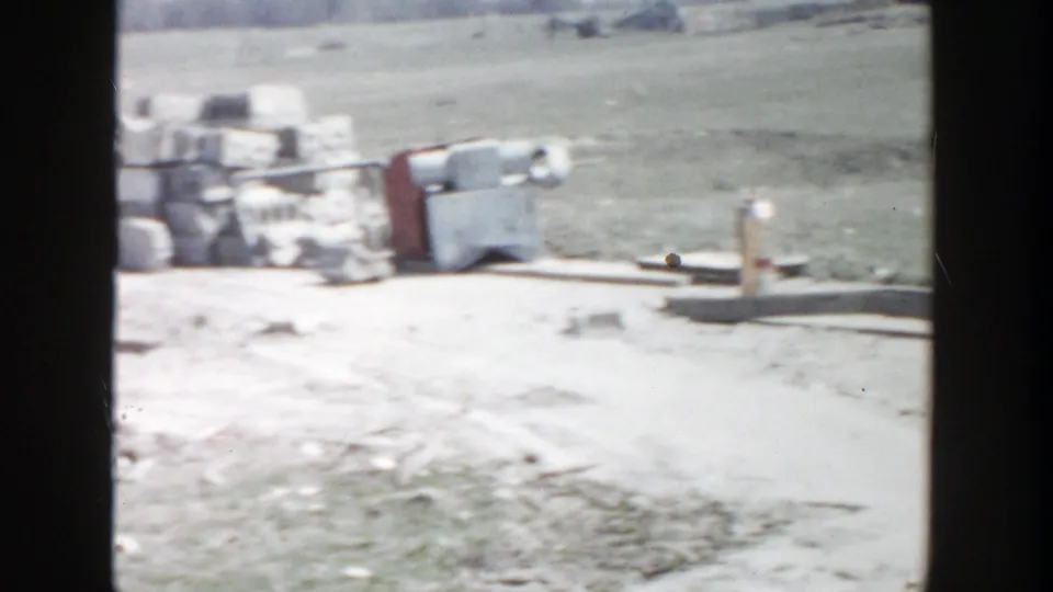 1956 Grand Rapids site view with stacked supplies, containers, open ground, and a small distant workman or figure near the materials.