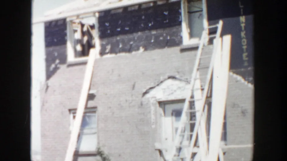 1956 worksite frame showing ladders on a brick building facade with open windows and visible sheathing at the upper wall.