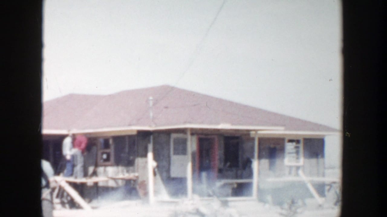 Workers stand near a partly built house with dust or smoke in the yard.