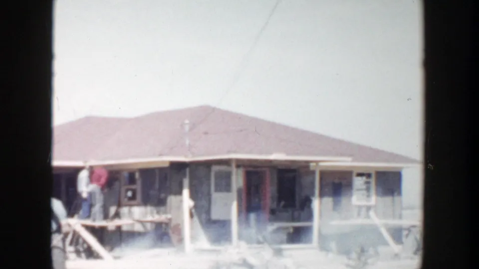 1956 Grand Rapids home-movie frame showing workers near a porch of a partly built house with pale dust or smoke across the yard.