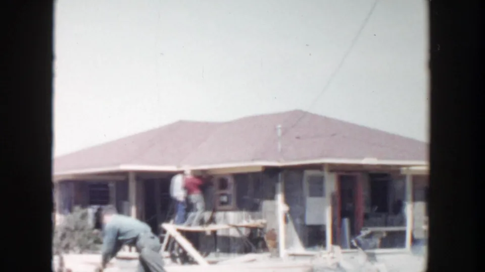A 1956 Grand Rapids yard scene shows one worker bending near the ground while others stand on the porch of a house under construction.