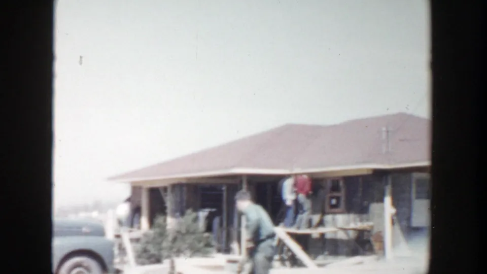 1956 Grand Rapids footage shows workers at the porch of a low house, with yard debris and construction activity visible in front.
