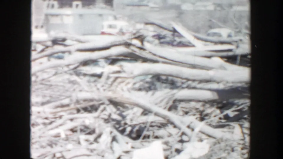 1956 Grand Rapids frame showing branches, a discarded tire, and pale debris with cars and low buildings in the background.