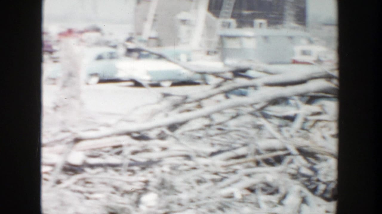 1956 Grand Rapids 8mm frame showing stacked branches or lumber across a cleared site with distant vehicles and buildings.